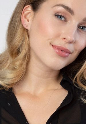 Gouden, delicate ketting met verfijnde schakels, gecombineerd met twee kleine studoorbellen. Het model heeft golvend blond haar en een heldere huid.