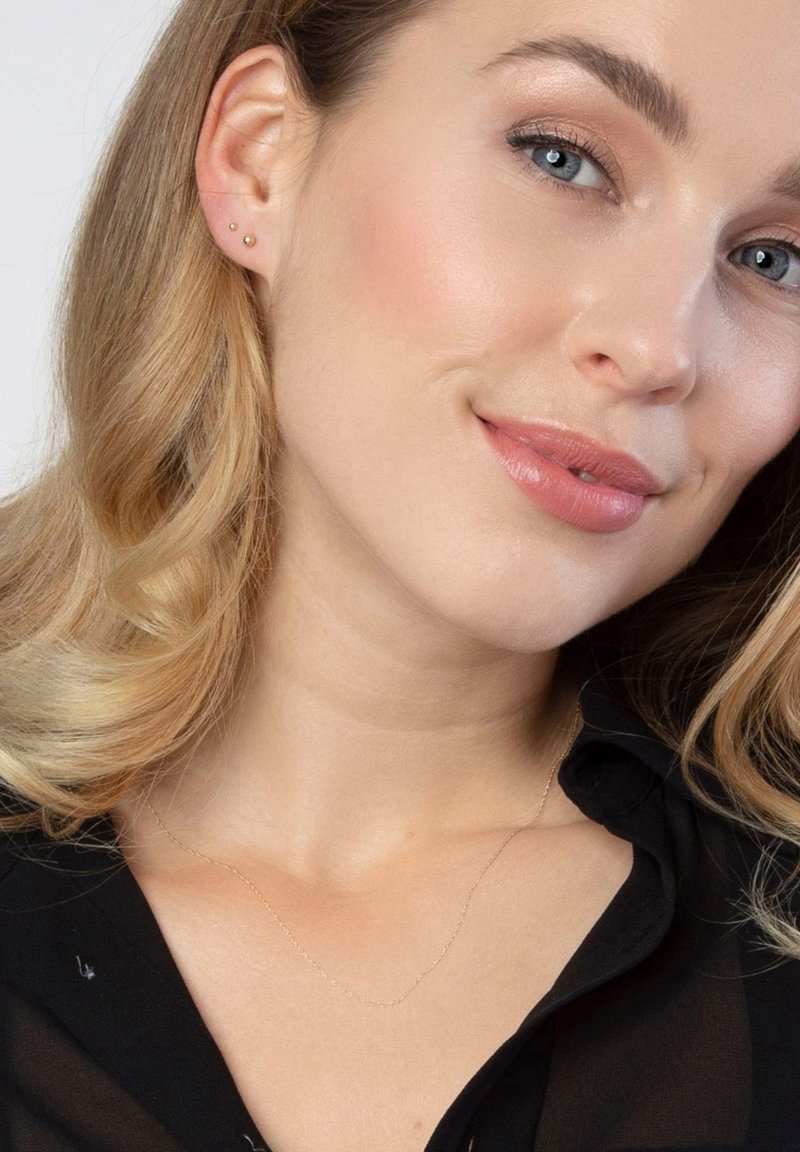 Gouden, delicate ketting met verfijnde schakels, gecombineerd met twee kleine studoorbellen. Het model heeft golvend blond haar en een heldere huid.