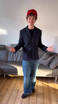 Blazer nero con due tasche frontali, indossato sopra una camicia nera, abbinato a jeans azzurri e scarpe nere. Un cappello rosso acceso completa l'outfit.