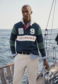 Marineblauw en groene rugbyshirt met witte strepen, grote "9" en logo patch, gelaagd over een blauw en wit gestreept shirt; beige broek.