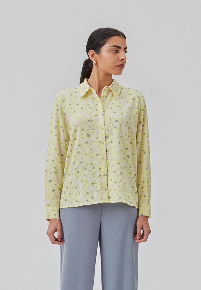 Gul skjorte med knaplukning i en tekstureret stof med små grå og lilla prikker, parret med lysegrå bukser.