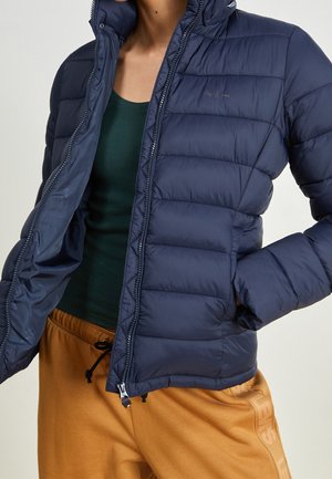 Osoba nosząca rozpiętą granatową pikowaną kurtkę na zielonym topie i musztardowych spodniach ze sznurkiem.