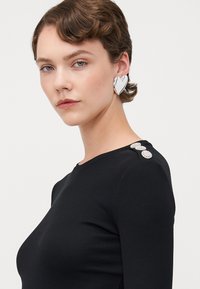 Black ribbed long-sleeve top featuring three decorative silver accents on the shoulder, paired with a large metallic heart-shaped earring.