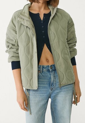 Personne portant une veste matelassée vert clair sur un haut bleu marine partiellement boutonné avec un jean taille haute bleu clair, montrant un ventre découvert.