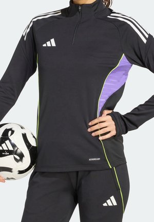 Personne en tenue de sport Adidas noire tenant un ballon de football noir et blanc, main sur la hanche, sur fond clair.