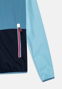 Jasnoniebieska i ciemnoniebieska wiatrówka z kieszenią na zamek, z kontrastowymi czerwonymi akcentami zamka błyskawicznego oraz gładką, lekko błyszczącą teksturą materiału.