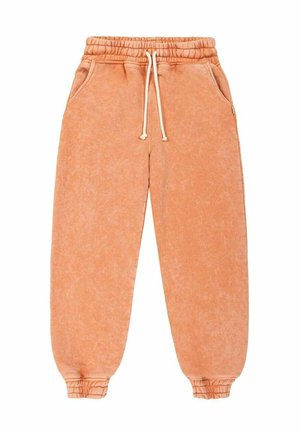 Osoba ubrana w brzoskwiniową bluzę i spodnie, na boso, stojąca na piaszczystej plaży z oceanem i pochmurnym niebem w tle.