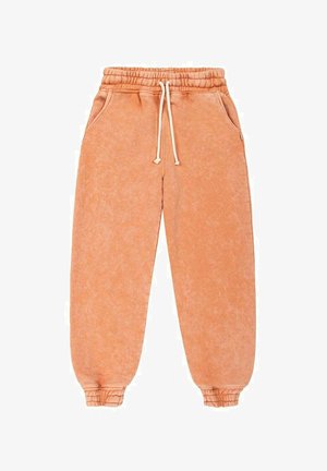 Person wearing peach-colored sweatshirt and pants, barefoot, standing on a sandy beach with ocean and cloudy sky in the background.