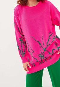 Oversized rosa tröja med grön abstrakt mönster, ribbad rundhals och manschetter, tillverkad av mjukt stickat material. Stylad med gröna byxor.