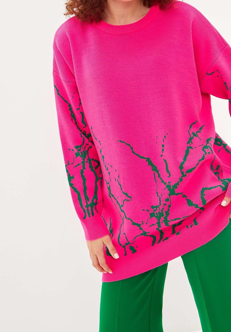 Oversized rosa tröja med grön abstrakt mönster, ribbad rundhals och manschetter, tillverkad av mjukt stickat material. Stylad med gröna byxor.