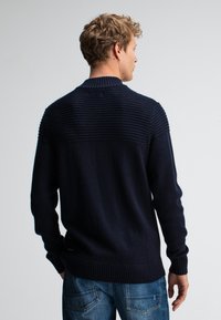 Man met kort, krullend haar, gekleed in een marineblauwe geribde trui en blauwe jeans, met zijn rug naar de camera tegen een effen lichte achtergrond.