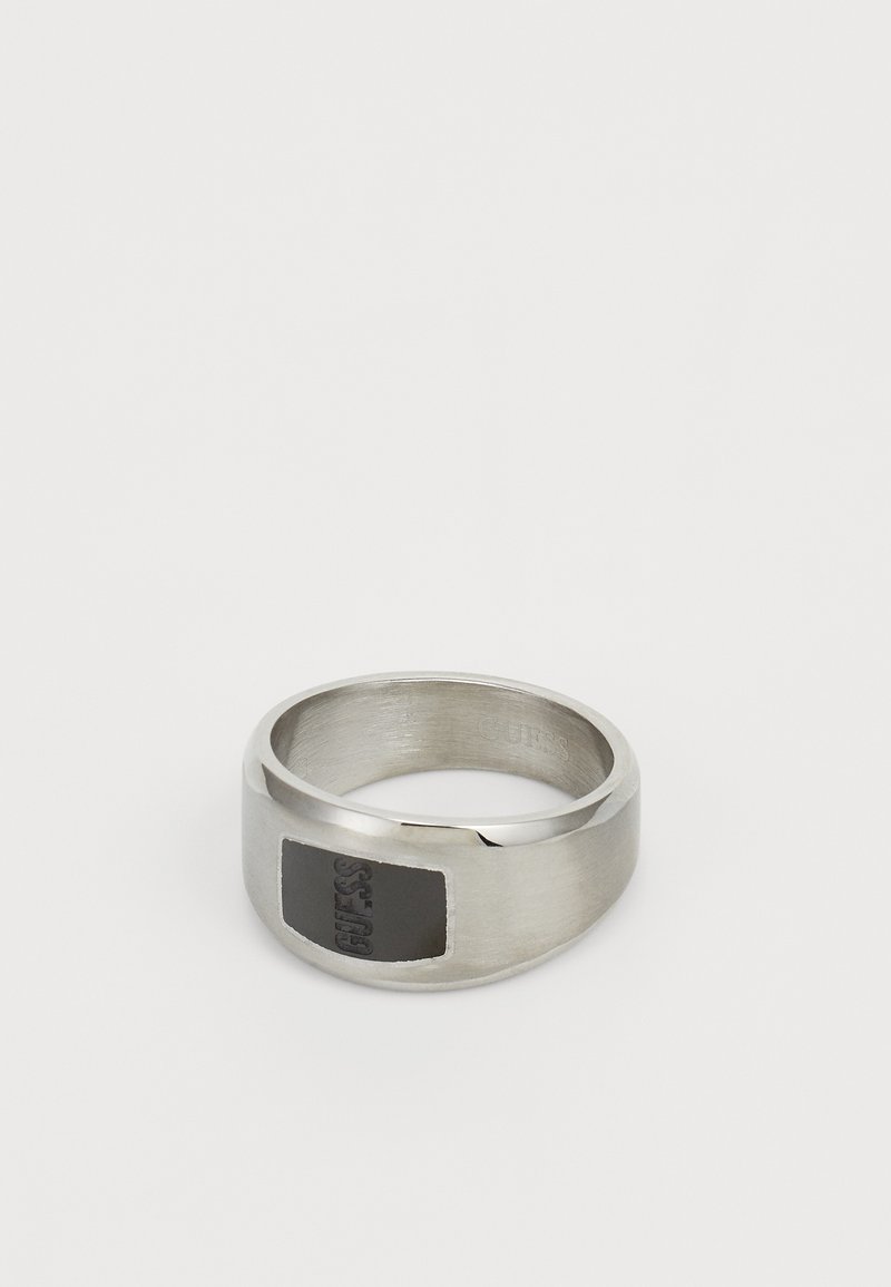 Bague en métal argenté avec un insert rectangulaire en émail noir portant la marque "GUESS" en relief. Surface lisse et polie avec des bords arrondis.