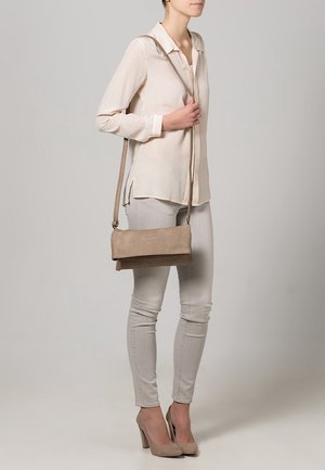 Bolso beige para el hombro con textura suave y diseño de solapa plegable. Lleva cruzado con una correa larga, combinado con un atuendo de tonos claros.