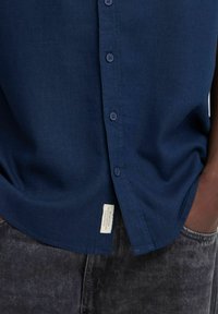 Námornícky modrá košeľa s gombíkmi vyrobená z texturovanej látky, s rovný strihom, šiestimi gombíkmi a malou nášivkou na leme.