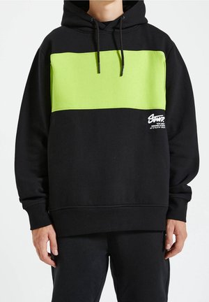 Osoba ubrana w czarną bluzę z kapturem z szerokim neonowozielonym poziomym paskiem i białym napisem na klatce piersiowej, w połączeniu z czarnymi spodniami.