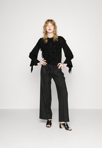 Blusa nera con volant e maniche lunghe, abbinata a pantaloni neri larghi e scarpe aperte nere. Sfondo da studio di colore neutro.