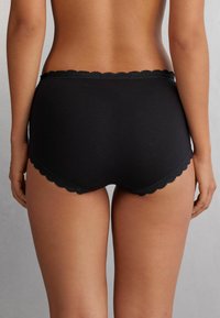 Svart bomulls hipsterunderkläder med vågig spetskant runt midjan och benen. Slät textur utan synliga mönster eller hårdvara.