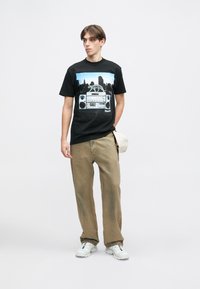 Melna kokvilnas T-kreklu ar boomboxa un ainavas grafiku. Apvienots ar bežām, brīvā stilā veidotām biksēm un baltām kedām.