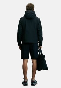 Zwarte sportjack met capuchon, gecombineerd met marineblauwe shorts. De persoon draagt een zwarte tas en heeft grijze sneakers met accenten aan.