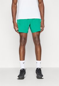 Grüne Shorts aus leichtem Material, mit Fronttaschen und figurbetontem Schnitt, kombiniert mit schwarzen Sneakers und weißen Socken.
