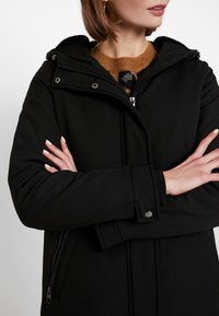 Chaqueta negra de softshell con capucha, que cuenta con bolsillos con cremallera y botones a presión, tela texturizada y un ajuste ceñido.