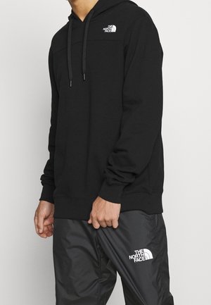 Mann trägt eine schwarze The North Face Kapuzenjacke und schwarze Hose mit weißem Logo, steht vor einem schlichten hellen Hintergrund.
