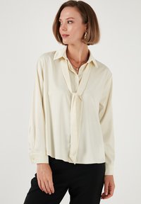 Blusa de manga larga color crema con collar y lazo en el escote, con una textura suave y un corte relajado.