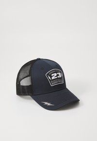 Tamsiai mėlyna kepurė su išlenktu kraštu, tinkleliu gale ir išsiuvinėtu logo, kuriame yra "Jordan Brand 23 Basketball Co." ir žaibo akcentai.