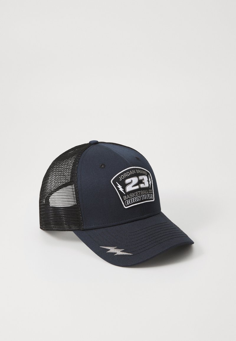 Tamsiai mėlyna kepurė su išlenktu kraštu, tinkleliu gale ir išsiuvinėtu logo, kuriame yra "Jordan Brand 23 Basketball Co." ir žaibo akcentai.