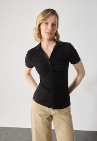 Black ribbed short-sleeve shirt with a fitted design, button-down front, and pleated detail at the bust, paired with light beige pants.