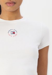 Camiseta de algodón blanca con mangas cortas, que presenta un logo multicolor de "tommy jeans New York" en un diseño circular en el pecho.