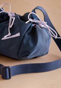 Tmavomodrá látková taška so zaťahovacím otvorom, vybavená kontrastnými svetlými šnúrkami a textúrovaným popruhom. Hladký povrch s jemným leskom.