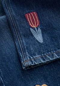 Denimstof in donkerblauw heeft kleurrijke borduursels van een rode tulp met gele accenten en groene bladeren. Bruine stiksels voegen detail toe.