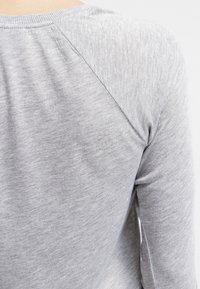 Graues Langarmshirt aus weichem, strukturiertem Stoff mit einem runden Ausschnitt und Raglanärmeln, die Nähte an den Schultern zeigen.