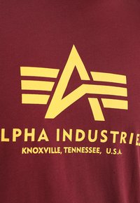 Mørkerød stof t-shirt med et stort gult stiliseret logo med et "A" og tre vandrette striber, mærket med "Alpha Industries."