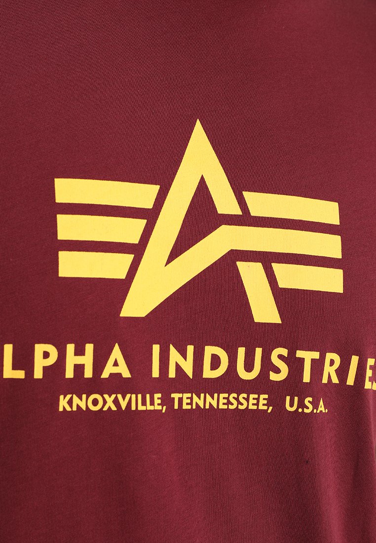 Maroonowa koszulka z tkaniny z dużym żółtym stylizowanym logo z literą "A" oraz trzema poziomymi paskami, oznaczona napisem "Alpha Industries".