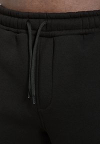 Černé tepláky s elastickým pasem a stahovací šňůrkou, boční kapsy a hladký povrch. Mají kontrastní stehy a kovové konce na stahovací šňůrce.