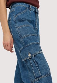 Modré denimové cargo kalhoty s několika prošívanými panely, boční kapsou se zapínáním na knoflík a vysokým pasem. Texturovaná tkanina a rezavě zbarvené švy.