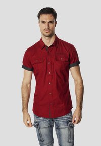 Camisa roja de manga corta con cierre de botones al frente, dos bolsillos en el pecho y puños remangados. Presenta tela texturizada y un sutil detalle de marca.