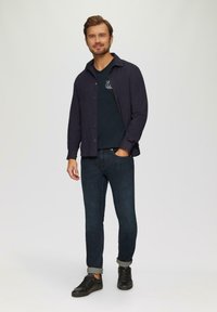 Navy knopshirt over een marineblauw T-shirt, gecombineerd met donkere denimjeans en zwarte sneakers. Heeft opgerolde mouwen en een eenvoudig ontwerp.
