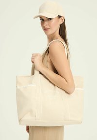 Beżowa torba na ramię z płótna z dwoma paskami na ramię, białym przeszyciem i przednią kieszenią z napisem "MELBOURNE TENNIS CLUB".