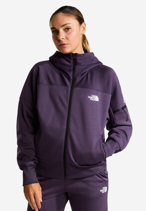 Kvinde iført en lilla hættetrøje med lynlås og matchende bukser med The North Face-logoet, hånd i lommen, kigger fremad med neutralt udtryk.