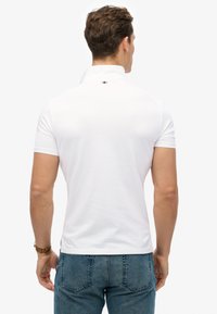 Biała koszulka polo z krótkim rękawem i kołnierzykiem; posiada mały kolorowy akcent na karku i wykonana jest z gładkiego, lekkiego materiału.