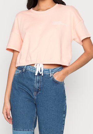 Femme portant un t-shirt court rose clair avec taille à cordon et un jean bleu taille haute, main dans la poche.