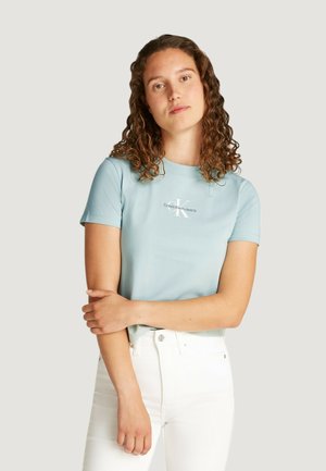 T-shirt in cotone azzurro chiaro con maniche corte che presenta un logo bianco di Calvin Klein. Il modello indossa dei jeans bianchi, con le braccia incrociate davanti.