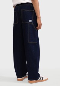 Wide-leg dark blue denim trousers with two large back pockets and contrasting yellow stitching. Paired with white sneakers.