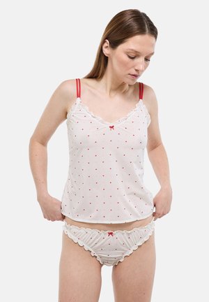 Femme portant un ensemble de lingerie blanche avec de petits cœurs rouges, des bretelles rouges et des nœuds, ajustant le bas du vêtement en regardant vers le bas.