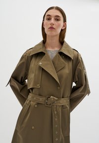 Trench coat lungo color khaki con vita cintata, ampio colletto e bottoni decorativi. Presenta maniche larghe e un tessuto liscio e testurizzato.