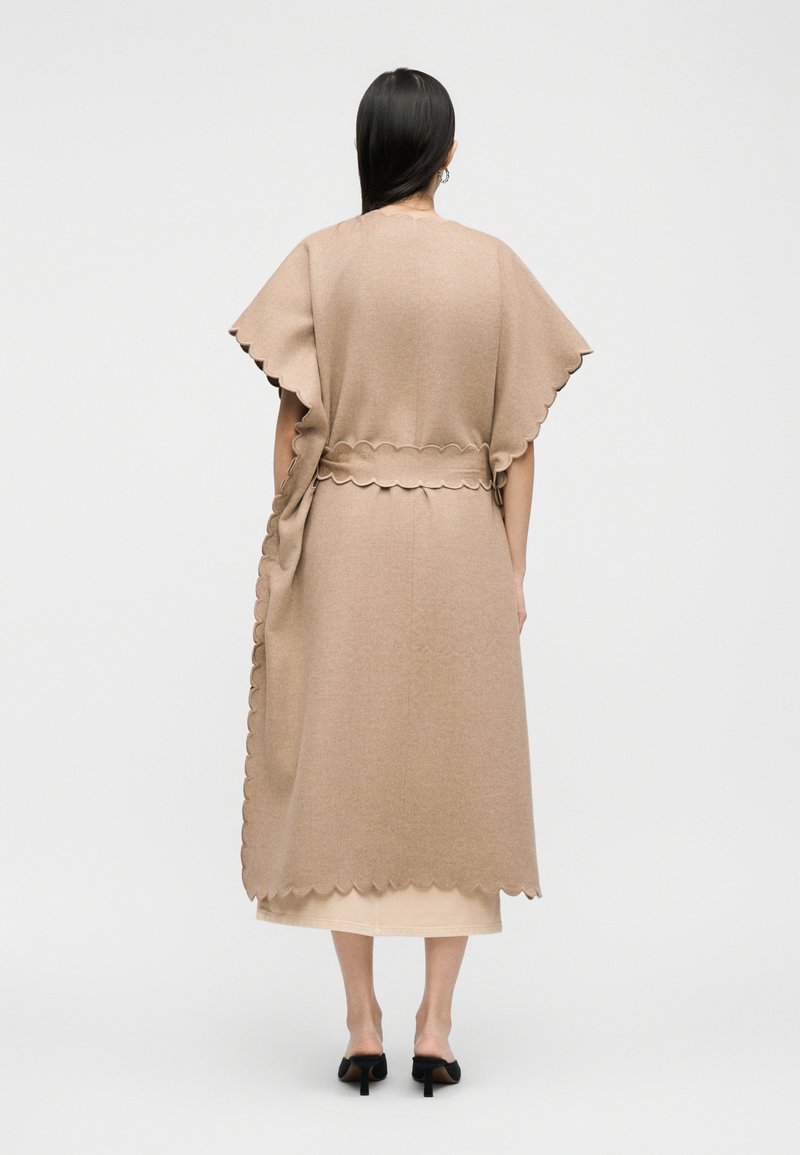 Frau mit langem dunklem Haar steht mit dem Rücken zur Kamera, trägt einen beigen Umhang mit wellenförmigem Saum und einen passenden Rock sowie schwarze Sandalen mit Absatz.