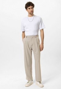 Wit t-shirt, beige geplooide broek en lichte sneakers. Model staat met handen in de zakken tegen een effen achtergrond.